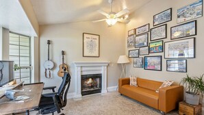 A living room with a fireplace, a sofa, a desk with a chair, a guitar, and framed artwork on the wall.