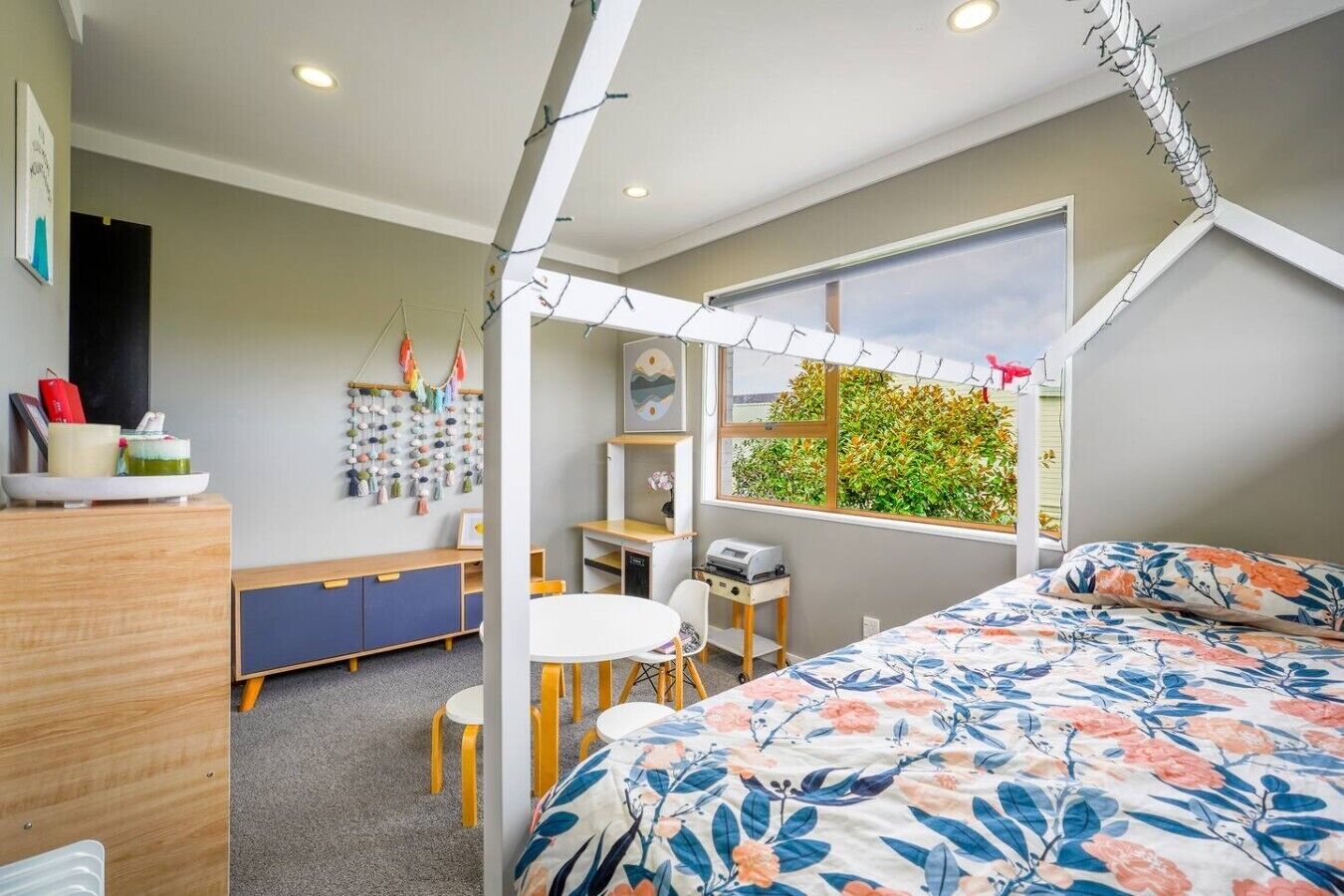 A bedroom with a sloped ceiling, a bed with a floral bedspread, a desk with a printer, a chair, and a window with a view of greenery.