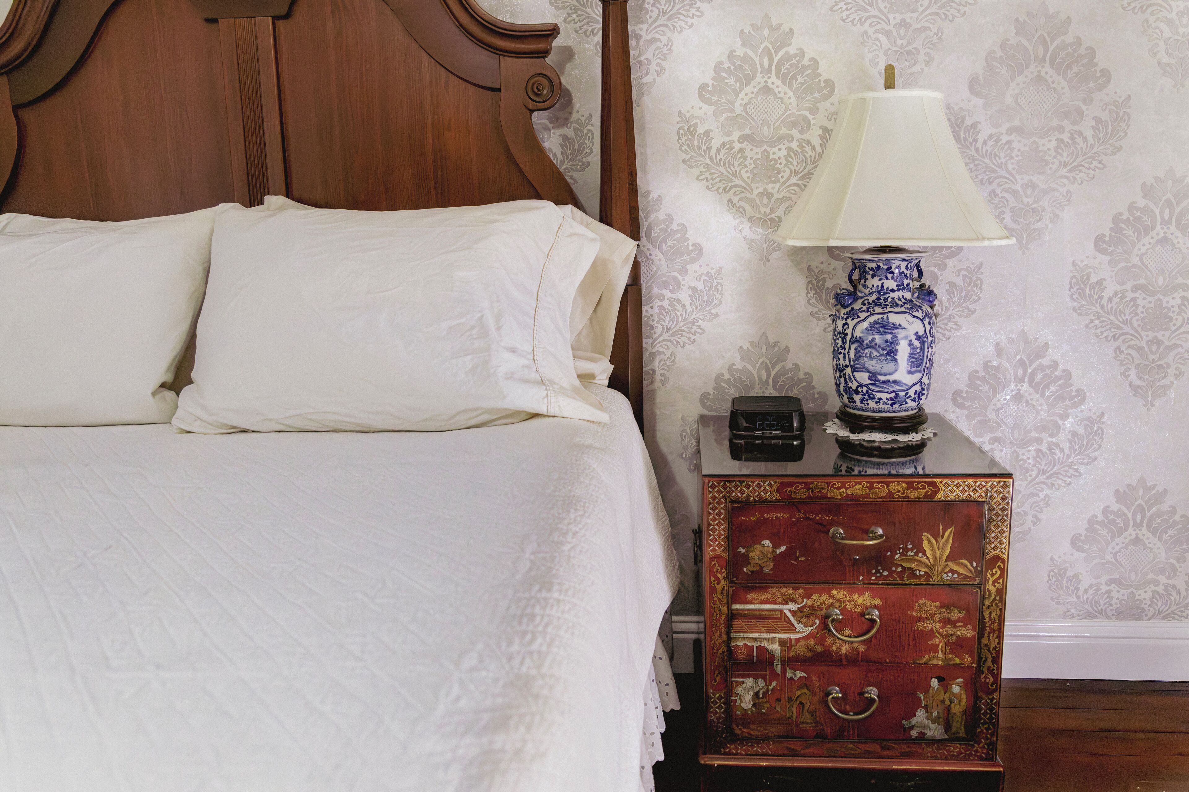 A neatly made bed with white linens, a wooden headboard, a decorative lamp, and a small red chest of drawers against a patterned wallpaper.