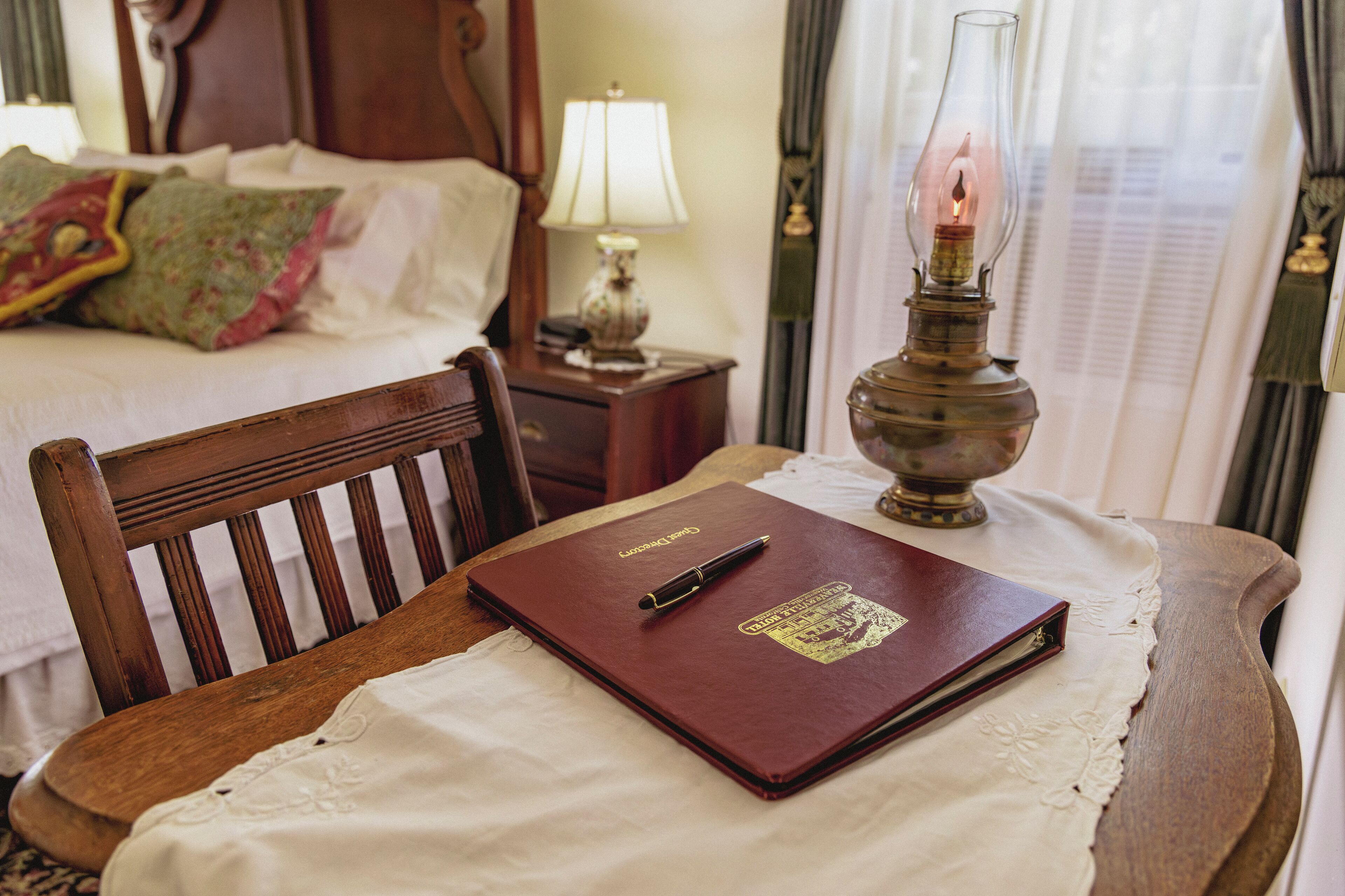 Uma cadeira de madeira com um caderno vermelho e caneta sobre uma mesa, uma lâmpada e uma cama ao fundo.