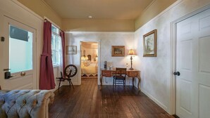 Un couloir avec un plancher en bois, un lit avec des rideaux rouges, un bureau avec une lampe et des tableaux encadrés accrochés au mur.