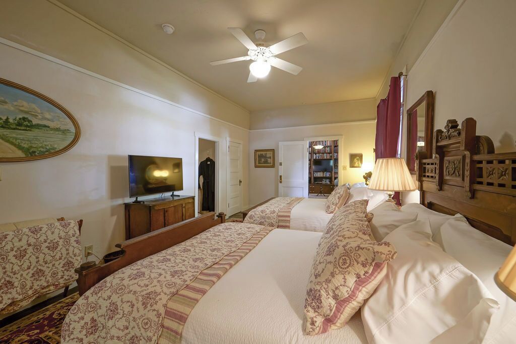 A hotel room with two beds, a television, a ceiling fan, and a bookshelf.