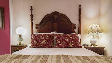 Cama bem arrumada com cabeceira de madeira, travesseiro floral e colcha listrada. Duas mesas de cabeceira com luminárias e um espelho do lado esquerdo.