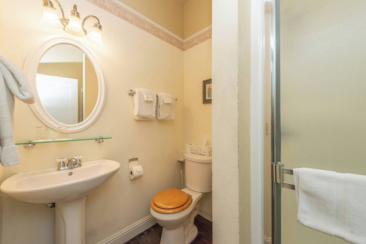 A bathroom with a pedestal sink, a toilet with a wooden seat, a glass shower enclosure, and a mirror with wall sconces.