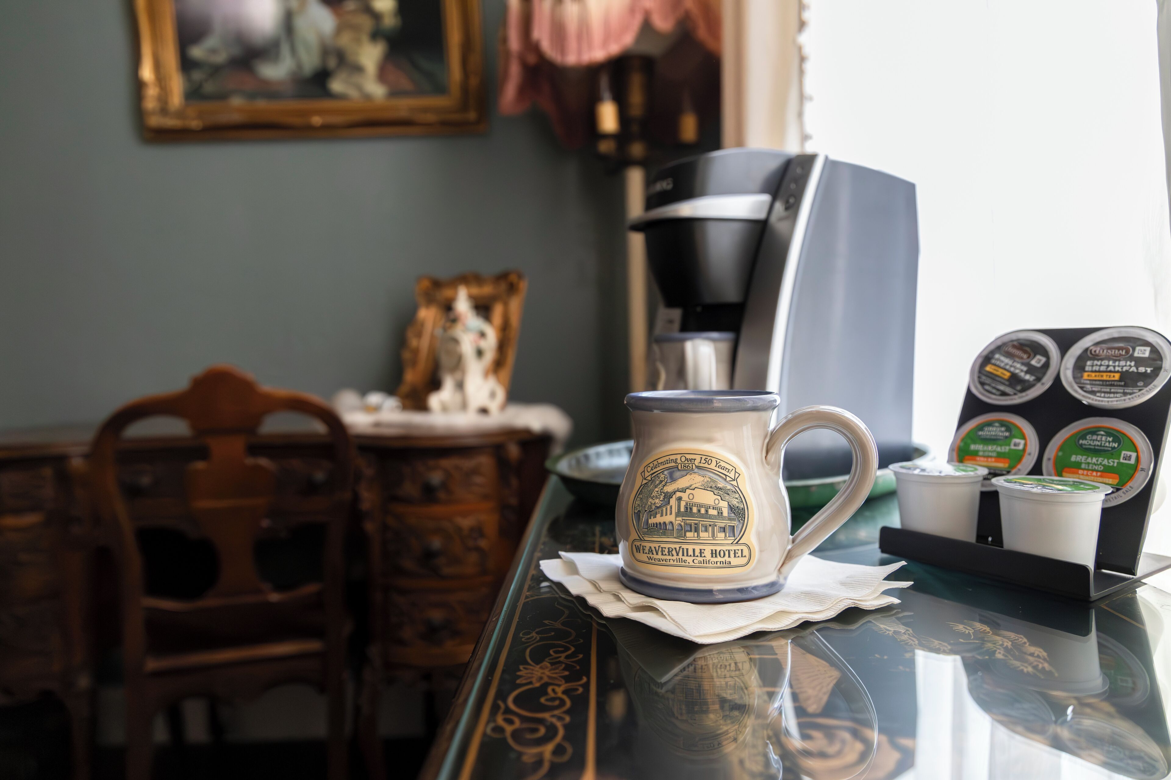 A coffee maker, a mug, and a coffee pod holder on a table with a decorative plate and a framed picture in the background.