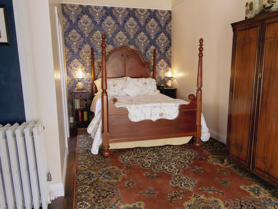 Una cama con dosel y tablero de madera, una alfombra estampada, un radiador y un armario de madera.