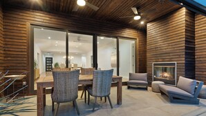 Un'area esterna moderna con zona pranzo, un tavolo in legno, sedie in vimini e una salottino imbottito, con un caminetto.
