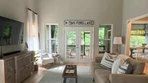 A spacious living room with a large television, a coffee table, and a sofa. There is a sign above the door that reads "TIMS FORD LAKE". - Rooted Lake Cottage w/ Golf Cart - NEW to VRBO! (Tullahoma)