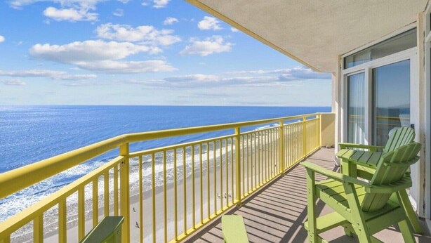 Un balcon avec une balustrade jaune, des chaises vertes et une vue sur l’océan.