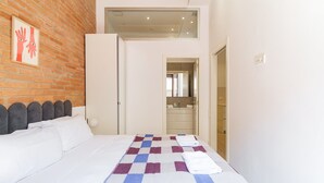 A hotel room with a bed, a checkered bedspread, a brick wall, a bathroom visible through an open door, and a small window above the bed. - GuestReady - Lovely home in the heart of Barcelona (Barcelona)