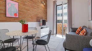 A modern living room with a brick wall, a dining area with a round table and chairs, a grey sofa with orange cushions, and a TV on a white cabinet. - GuestReady - Lovely home in the heart of Barcelona (Barcelona)