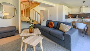 Una sala moderna con un sofá gris, una escalera de madera, un comedor con mesa y sillas, y una cocina al fondo.