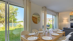 Comedor con una mesa de madera preparada para seis personas, sillas blancas y vista a un jardín a través de amplios ventanales correderos.