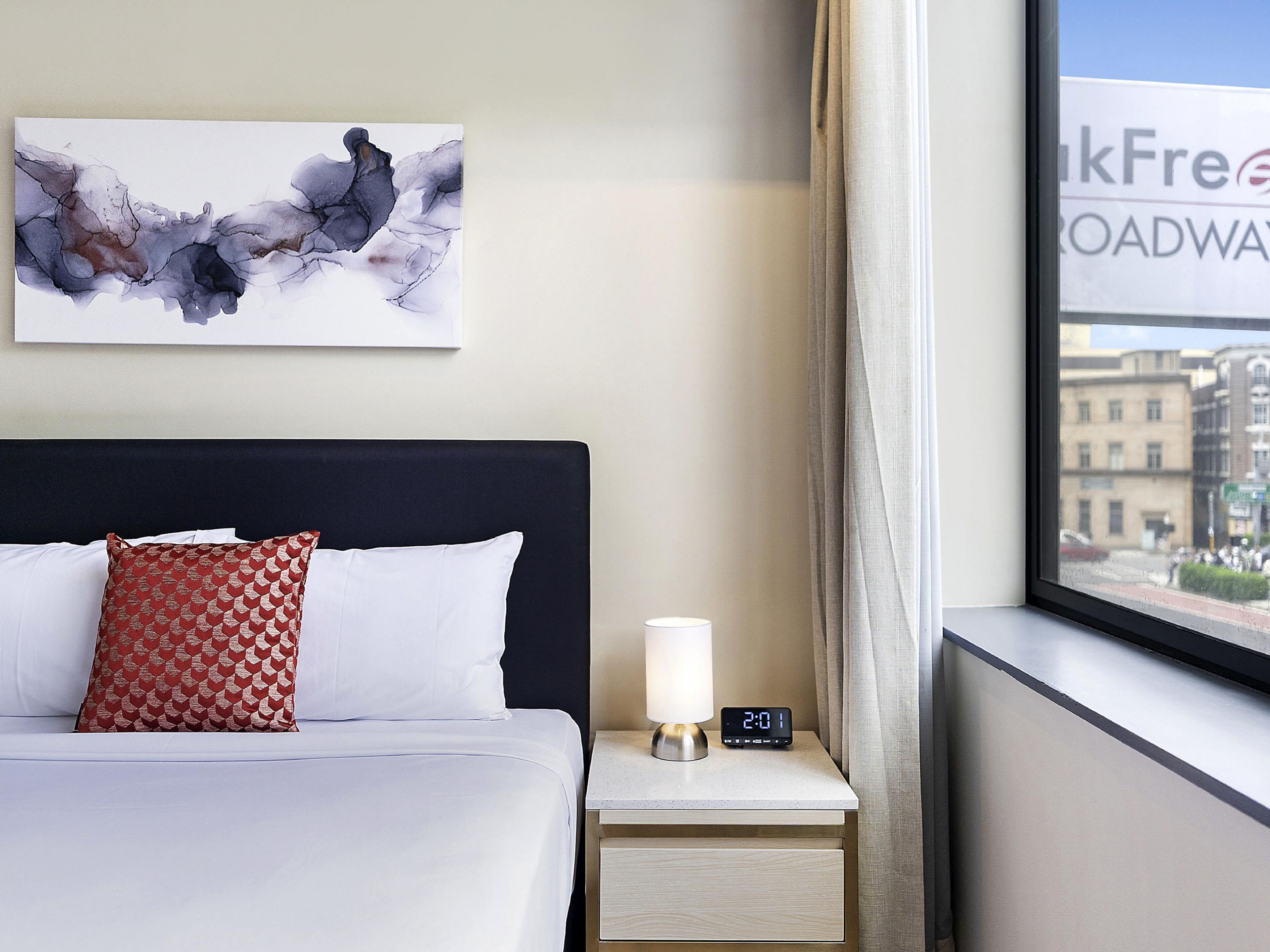 Et hotelværelse med en seng, et natbord med et ur, et vindue med udsigt til bygninger og et kunstværk på væggen.