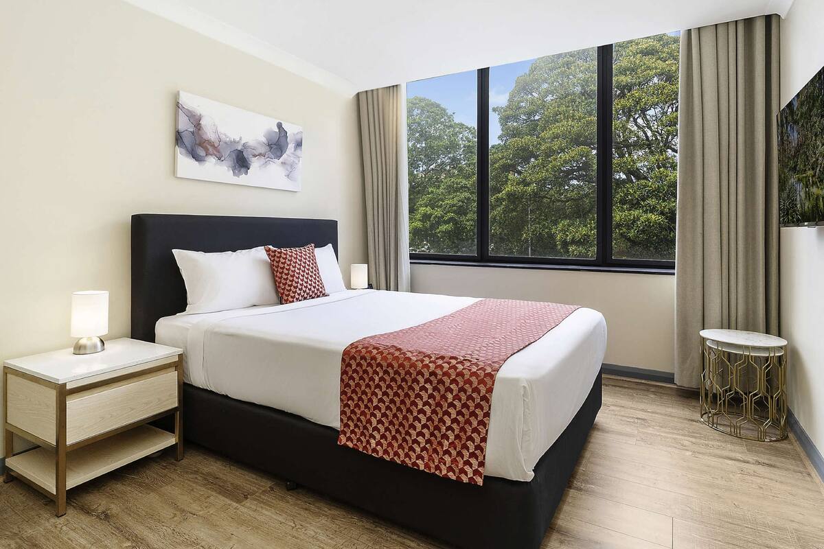 Une chambre d’hôtel avec un grand lit, une table de chevet, une commode, une fenêtre avec des rideaux et un tableau au mur.