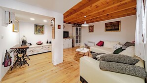 Ein gemütliches Wohnzimmer mit Holzbalkendecke, einem weißen Sofa mit Kissen, einem kleinen weißen Tisch und einer Nähmaschine in der Ecke.