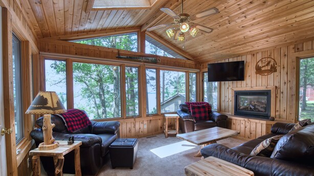 Interior de una cabaña de madera con chimenea, muebles de cuero y ventanas amplias.
