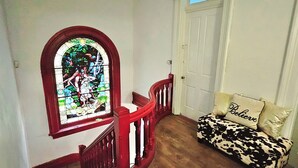 Un escalier orné d’une vitre en verre coloré, une rampe en bois et un canapé à motifs de vaches, agrémenté d’un coussin sur lequel est brodé le mot « Croire ».