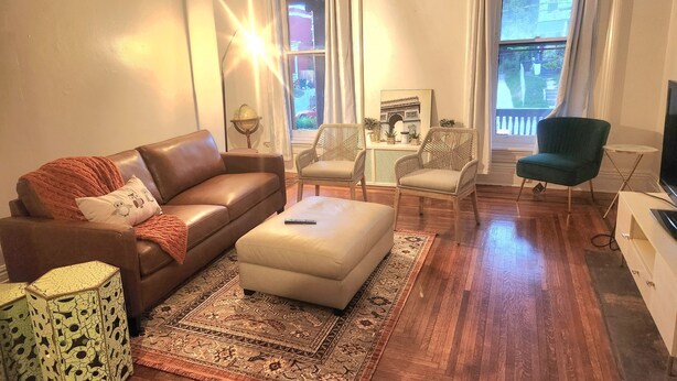 Un salon avec un canapé en cuir marron, un pouf beige, des chaises en osier, un fauteuil vert, une petite table blanche et une lampe en forme de globe.