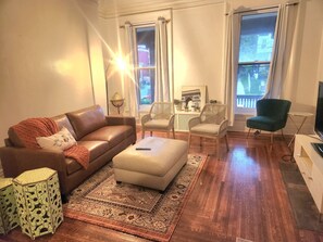 A living room with a brown leather sofa, a beige ottoman, wicker chairs, a green armchair, a small white table, and a globe lamp. - Comfortable 8-bdrm, 3-bath with off street parking, near XU, UC, OTR & Downtown (Cincinnati)