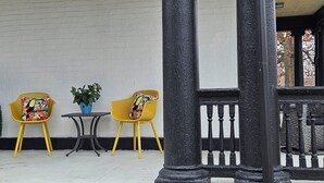 Un bâtiment blanc avec une balustrade noire, deux chaises jaunes ornées de coussins à motifs floraux, une petite table et une plante en pot.