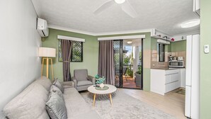 Una sala de estar con un sofá gris, una mesa de centro redonda y un sillón gris. Hay un ventilador de techo, un equipo de aire acondicionado y una zona de cocina con microondas y refrigerador.