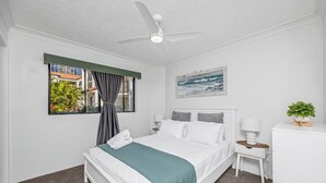 Una habitación de hotel con una cama, una mesita de noche, un escritorio, un ventilador de techo, una ventana con cortinas y un cuadro en la pared.