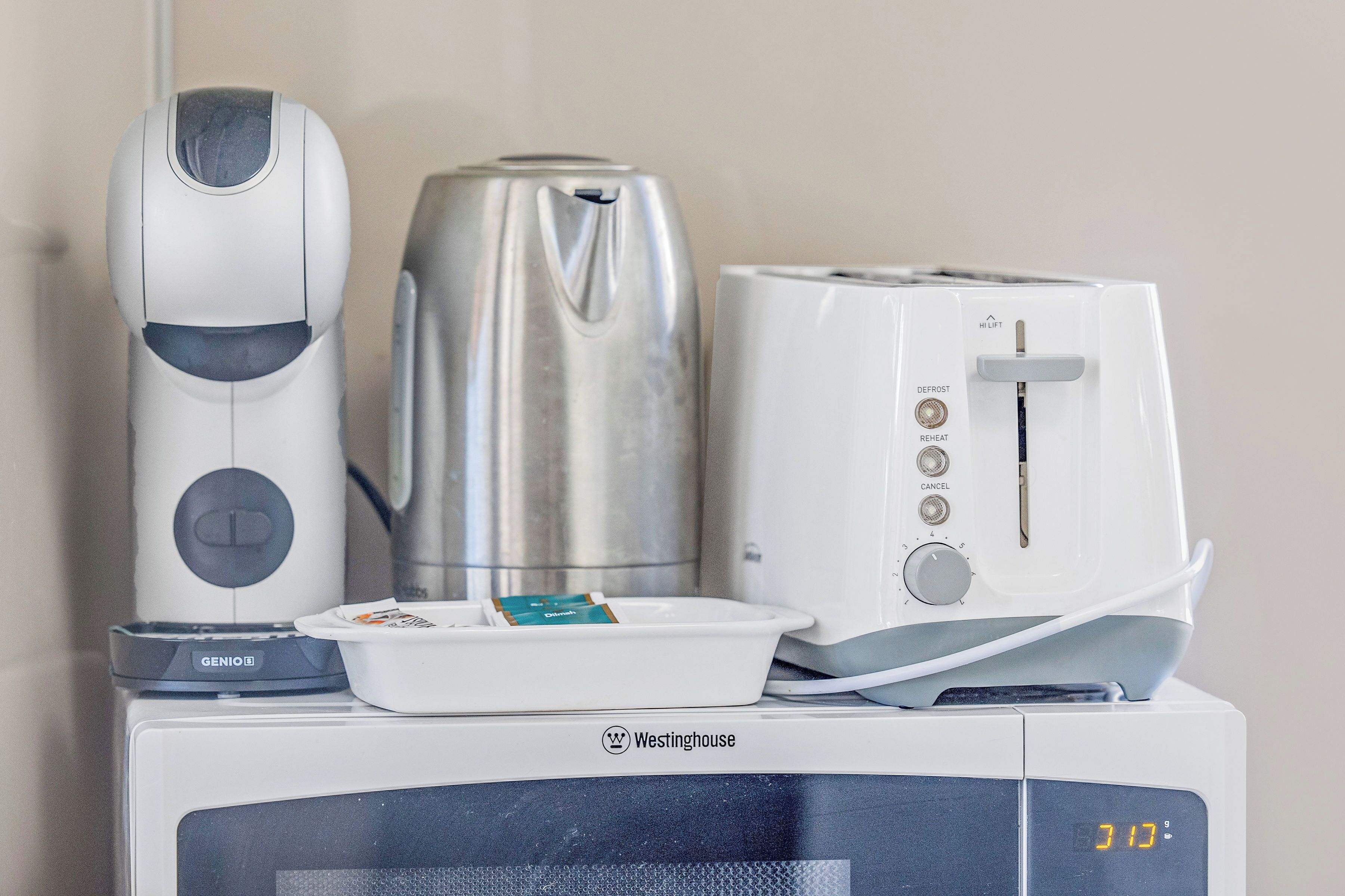 Um micro-ondas Westinghouse com relógio digital mostrando 3:10, uma cafeteira branca, uma chaleira prateada e uma torradeira branca, tudo sobreposto.