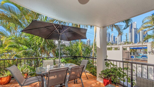 Un balcón con mesa, sillas y una sombrilla, con vistas a un paisaje urbano con palmeras.