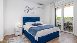 Une chambre moderne avec une tête de lit bleue, une table de chevet blanche, une commode, un grand lit avec une literie blanche et bleue, une citation décorative sur le mur et une vue sur la verdure à travers une porte-fenêtre coulissante.