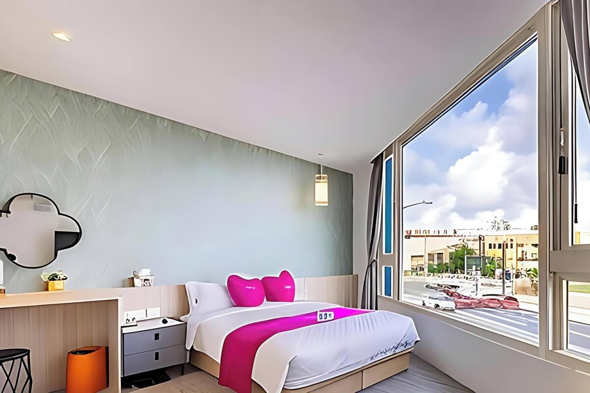 Quarto de hotel moderno com uma janela grande, uma cama com almofadas cor-de-rosa, uma escrivaninha com uma luminária e um espelho em formato de nuvem fixado na parede.
