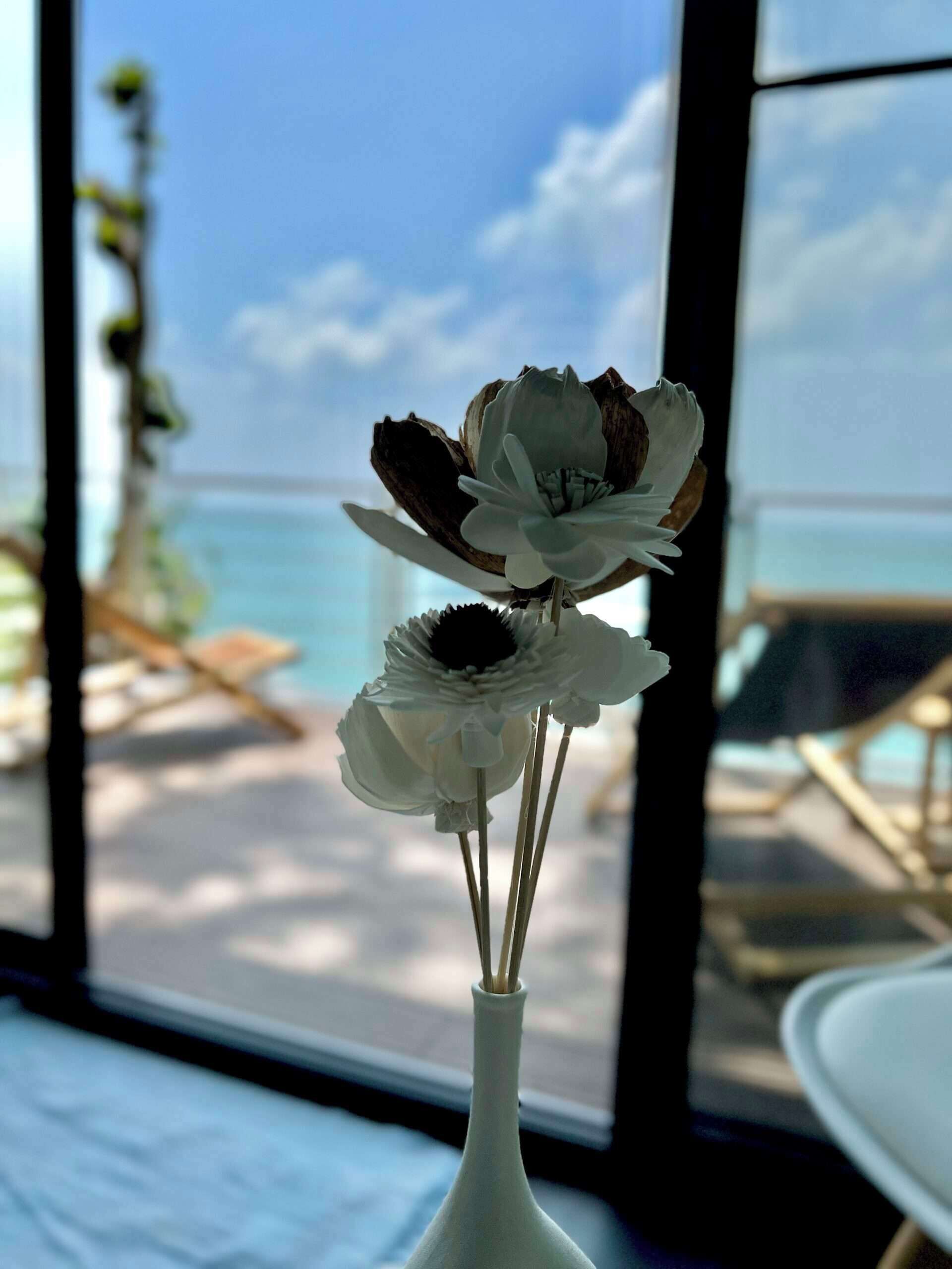 A white vase with flowers on a windowsill, overlooking a beach and lounge chairs.