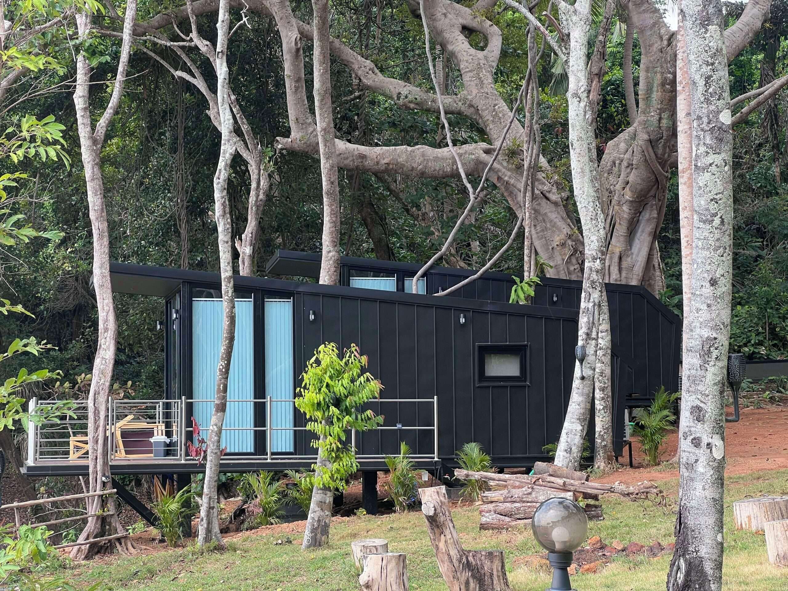 Una cabaña moderna negra con terraza y ventanas grandes, rodeada de árboles y follaje.