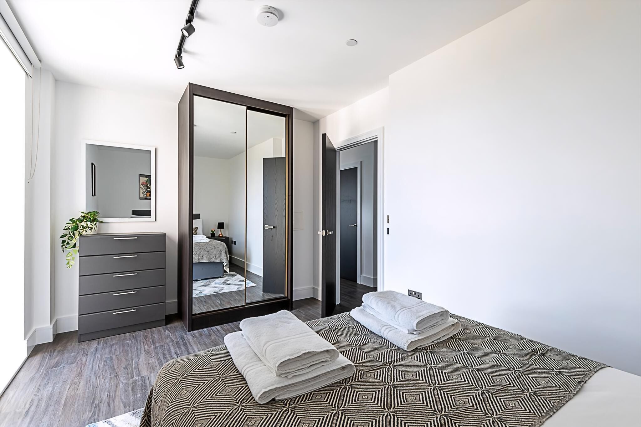 A modern bedroom with a large bed, a wardrobe, a mirror, and a plant.