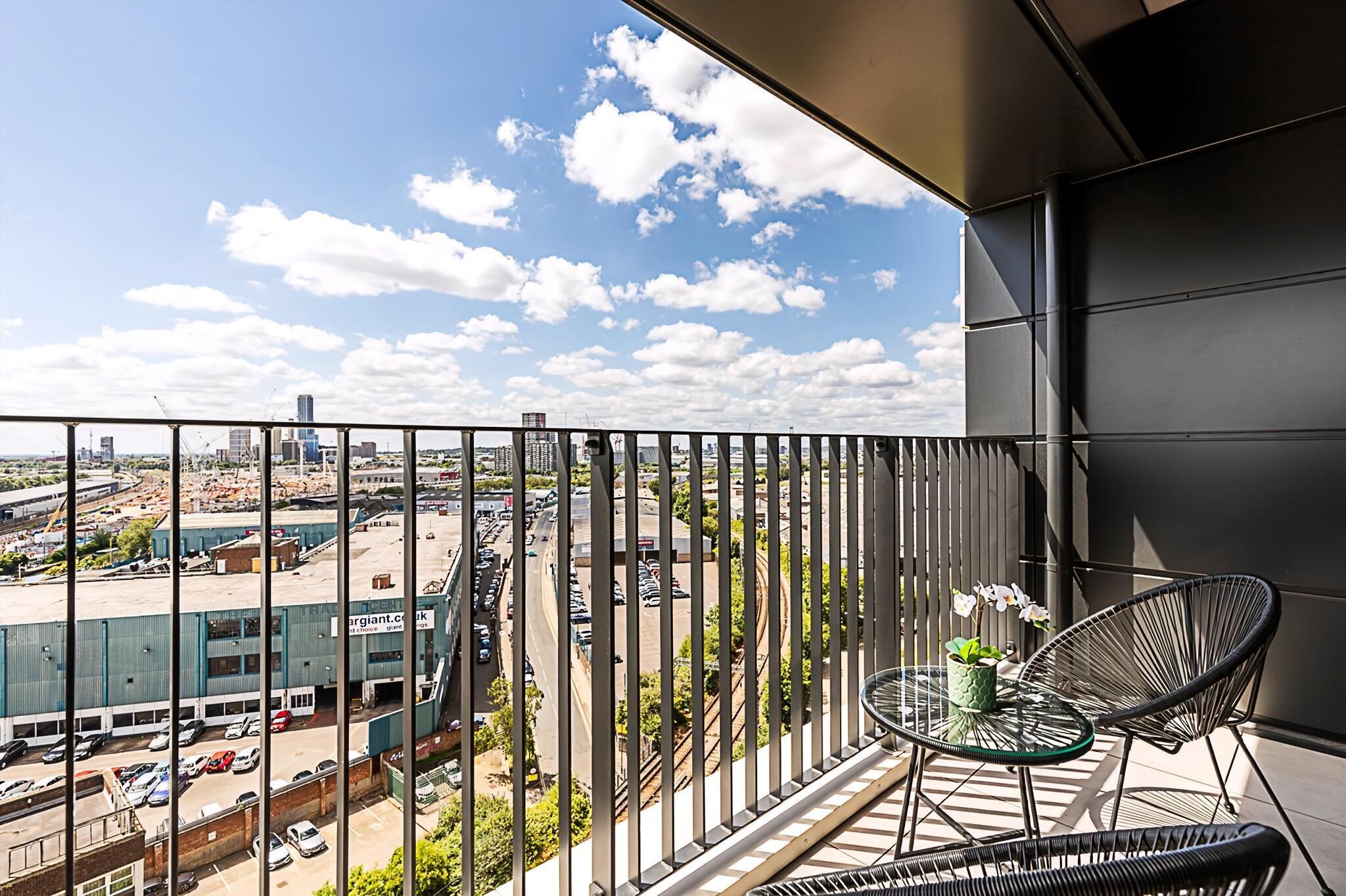A balcony with a view of a parking lot and buildings, a round table with a plant, and two wicker chairs.