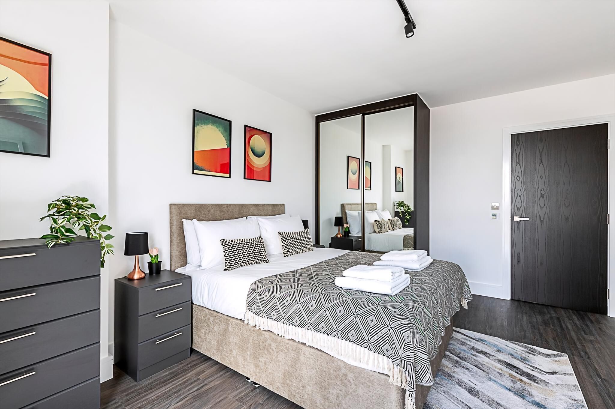 Un dormitorio moderno con una cama grande, un armario de madera oscura, una mesita de noche con lámpara y dos cuadros abstractos en la pared.