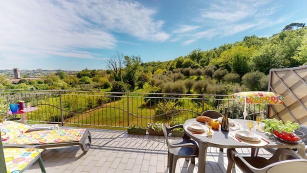 Una terraza con mesa lista para comer, sillas, un paraguas colorido y vistas a un paisaje verde exuberante.