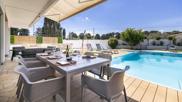Zona de piscina al aire libre con un patio cubierto, sillas de descanso y una mesa de comedor preparada para seis personas.