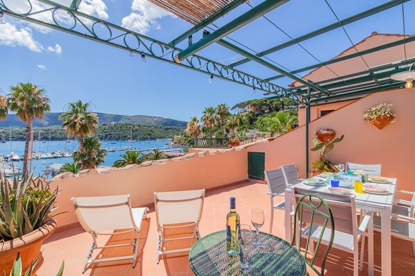 A terrace with a table set for dining, overlooking a marina with boats and a coastal landscape.