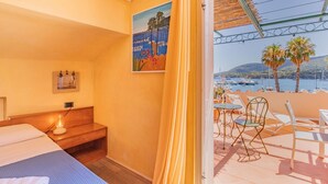 Quarto de hotel com cama, mesa de cabeceira com abajur, pintura na parede e varanda com mesa e cadeiras com vista para um calhavete.