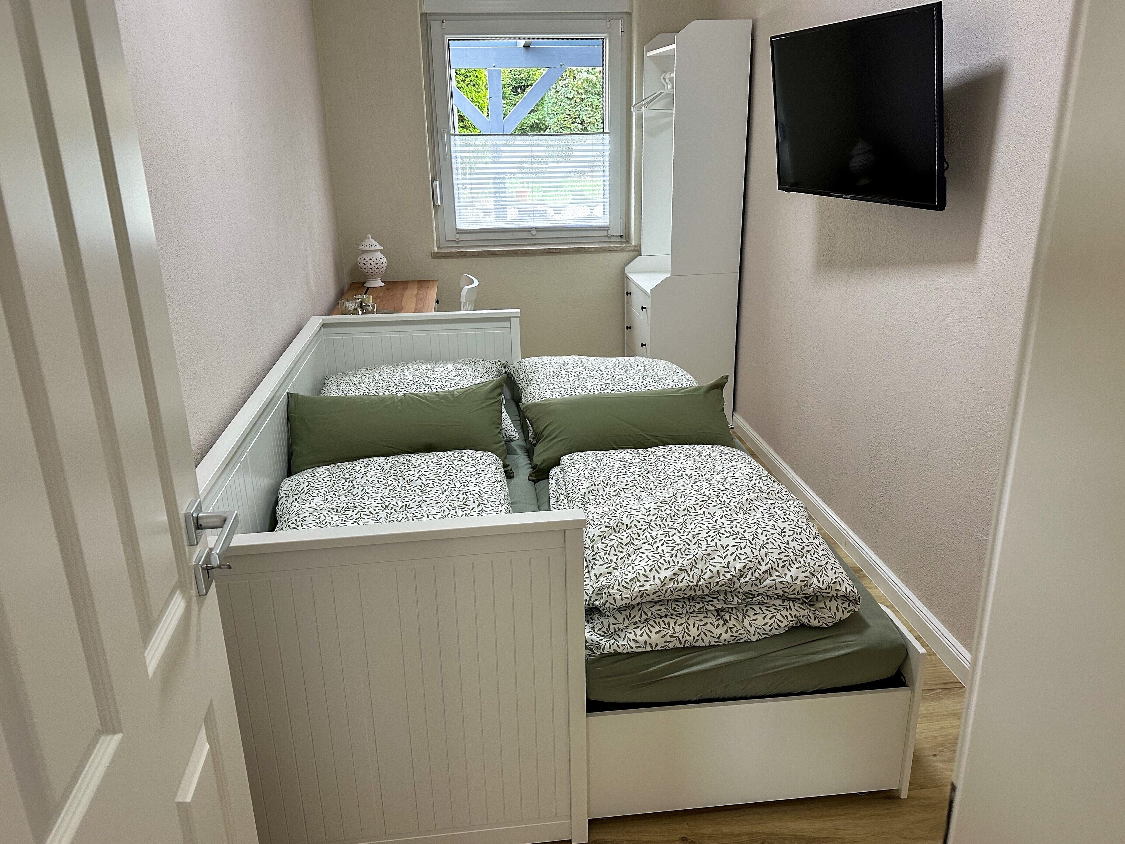 Ett smalt rum med två sängar, ett fönster med persienner, en tv på väggen och ett litet bord med en lampa.