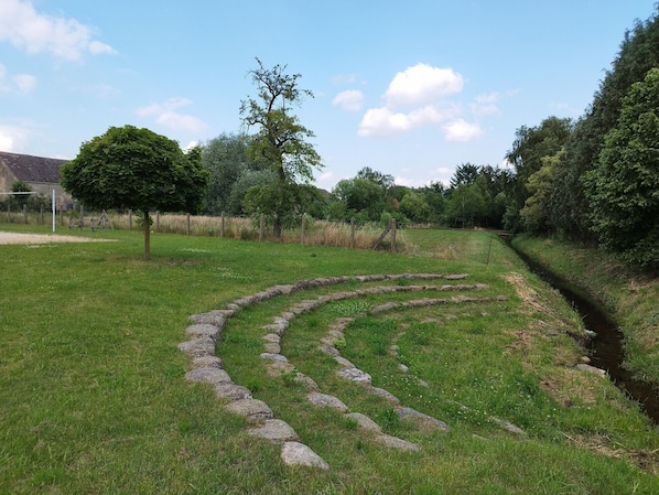 Un champ verdoyant traversé par un sentier de pierres qui serpente à travers l’herbe, bordé d’arbres et d’un petit ruisseau.