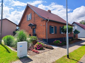 Une maison à deux étages, construite en briques, avec un toit recouvert de tuiles rouges, un petit porche à l’avant et un jardin agrémenté de fleurs et d’arbustes.