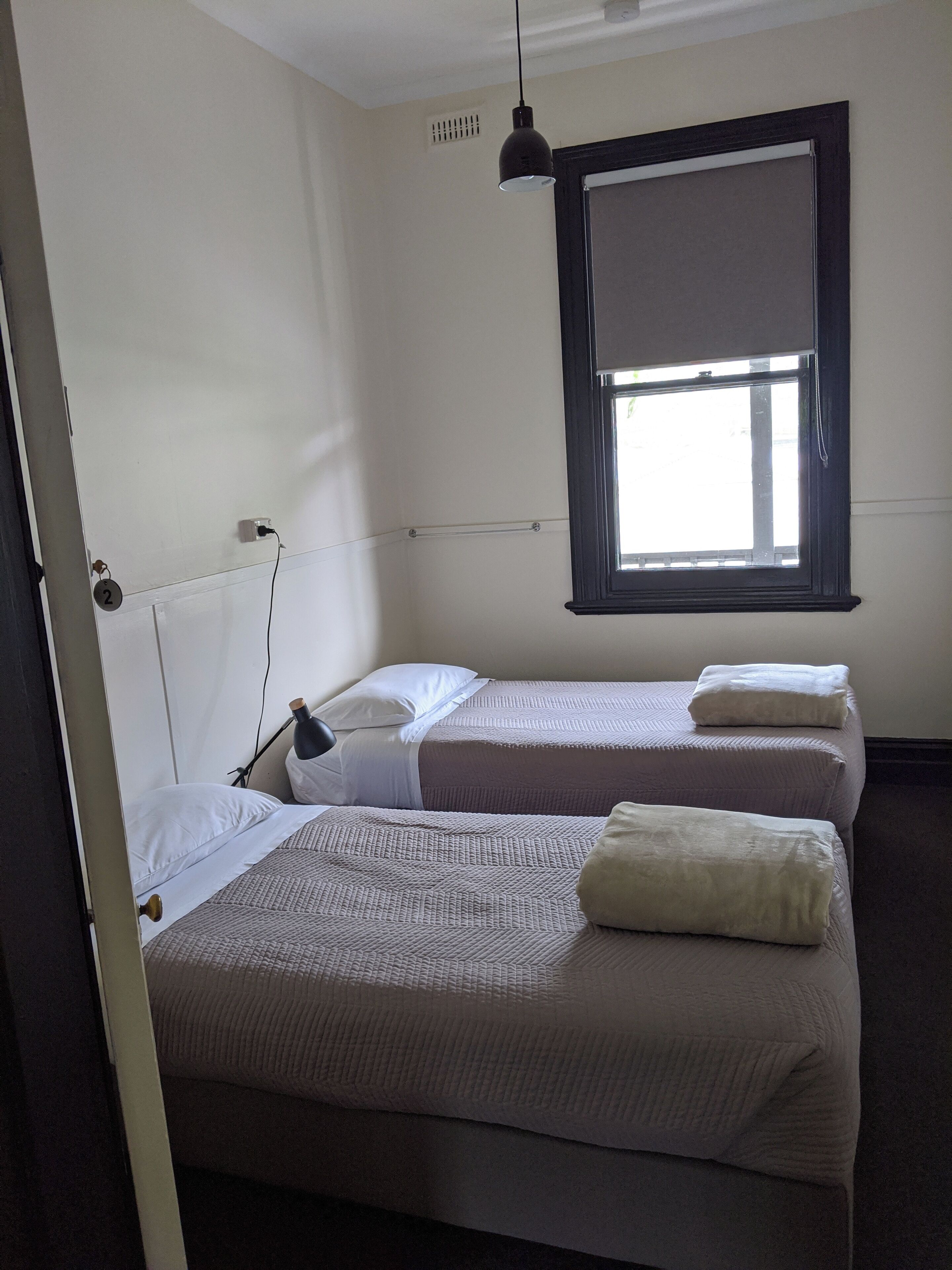 A room with two beds, a window with blinds, a hanging light fixture, and a visible electrical outlet.