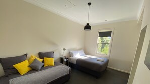 Een moderne slaapkamer met een slaapbank, een enkel bed, een klein tafeltje met een lamp en een raam met uitzicht op groen.
