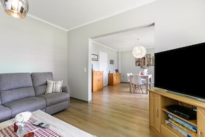 Una sala de estar con un sofá, una mesa de centro con un florero, un televisor de pantalla plana sobre un soporte de madera, y un comedor con sillas y una mesa.