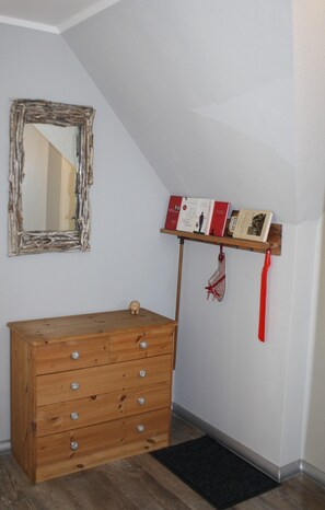 A wooden dresser with drawers, a mirror with a rustic frame, a shelf with books and a red ribbon, and a black doormat.