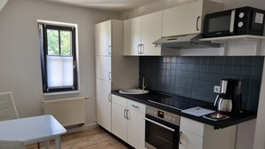 Kompakti keittiö, jossa on valkoiset kaapit, musta työtaso, mikroaaltouuni, kahvinkeitin ja ikkuna, jossa on sälekaihtimet.