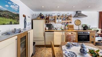 Een keuken met een stenen vloer, houten kastjes, een eettafel gedekt met blauw en wit servies, en een schilderij van een landschap aan de muur.