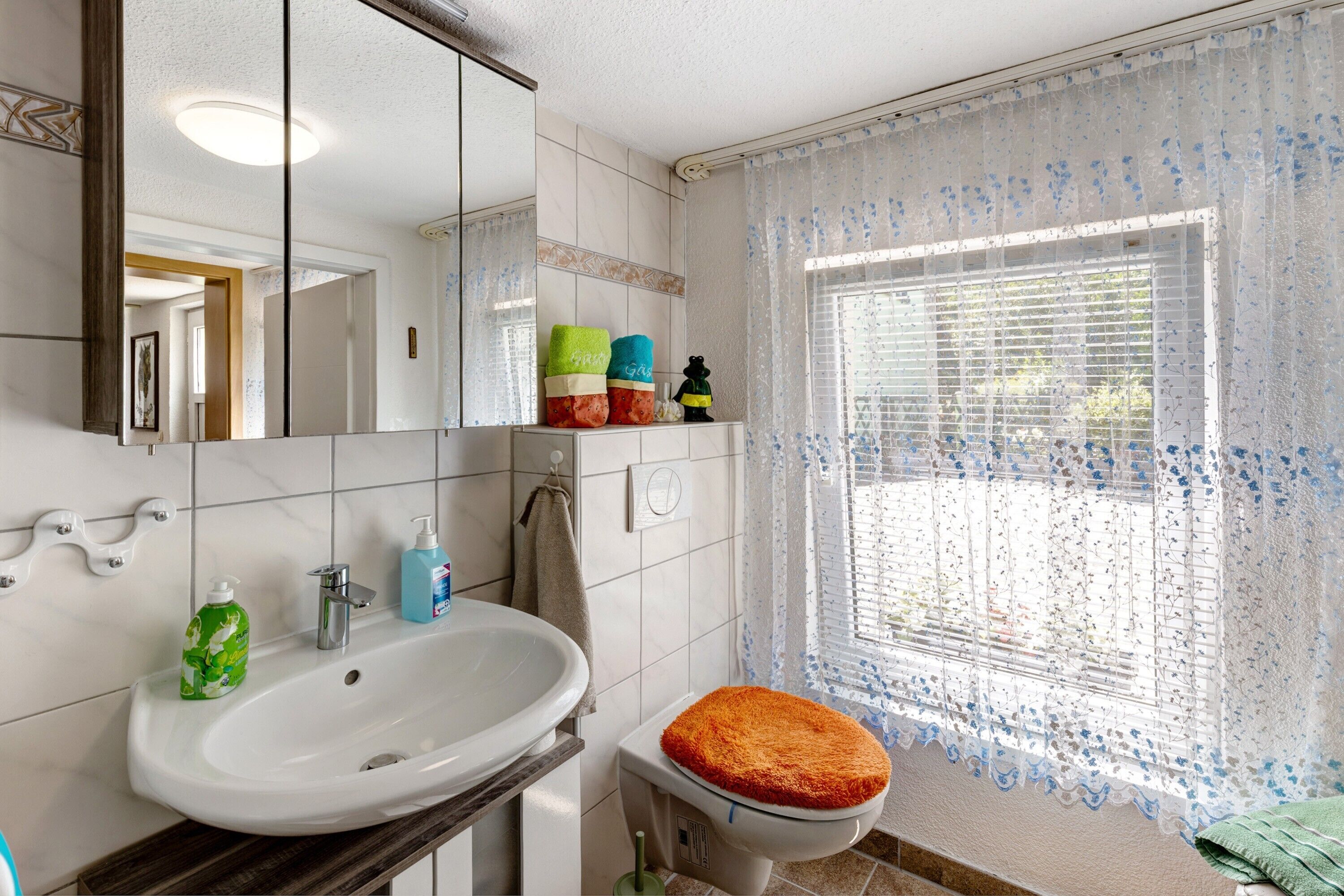 A bathroom with a white sink, mirror, and toilet. There are toiletries on the counter and a window with a patterned curtain.